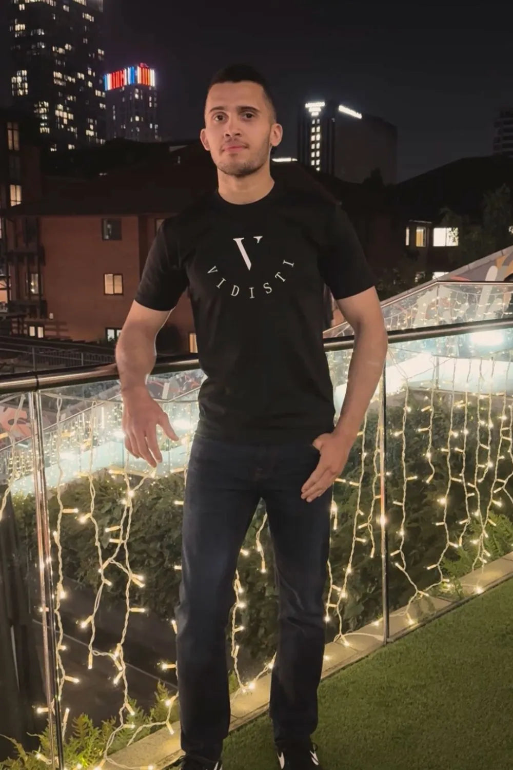 Man wearing a black t-shirt with a logo, standing on a balcony with decorative lights at night.