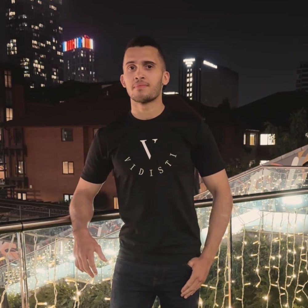 Man wearing a black t-shirt with a logo, standing on a balcony with decorative lights at night.