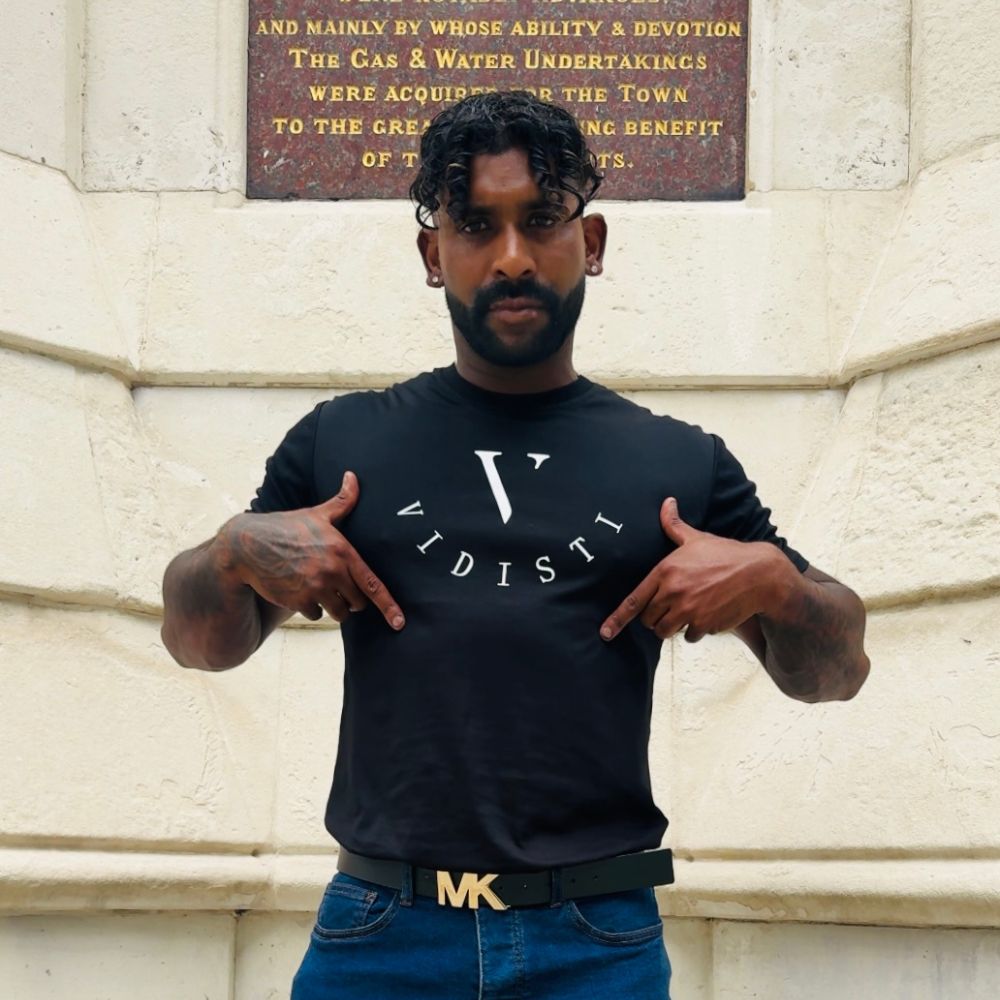 Person wearing a black t-shirt with a logo and blue jeans standing in front of a stone wall.