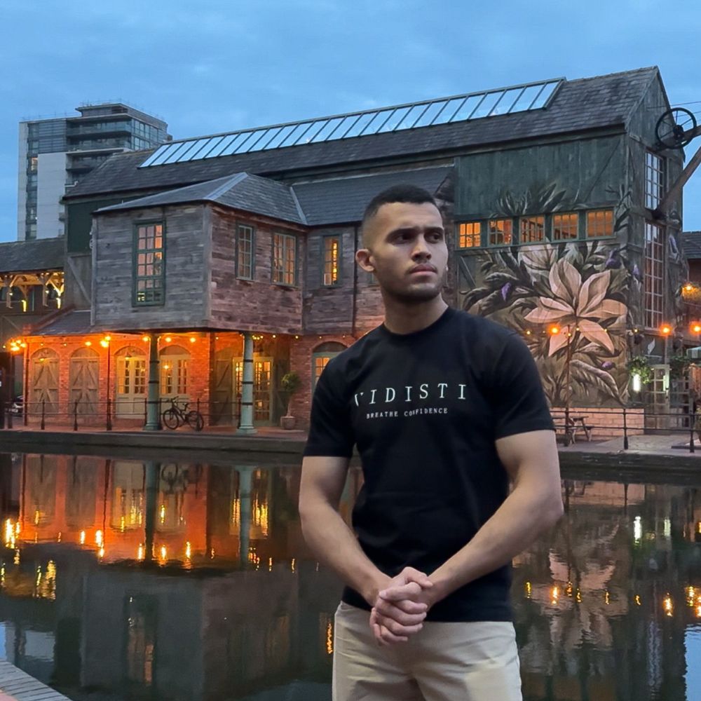 Man wearing a black t-shirt with 'VIDISTI' logo standing in front of a building with a mural.