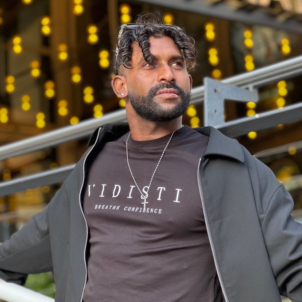 Man wearing a dark brown t-shirt with 'VIDISTI' text and a gray jacket, standing against a blurred background with lights.